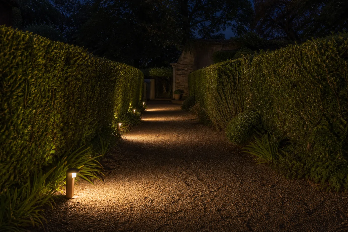 Allée de gravier éclairée par des bornes basses, lumière chaude vers le sol
